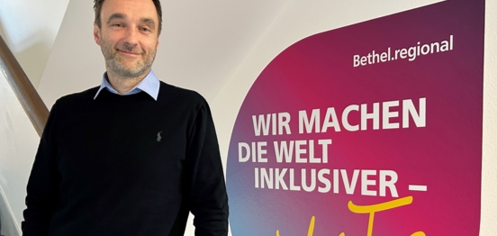 Auf dem Foto steht ein Mann an ein Geländer gelehnt, im Hintergrund an der Wand klebt der Spruch "Wir machen die Welt inklusiver - jeden Tag", Bethel.regional