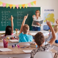 Mehrere Kinder sitzen in einem Klassenraum und werden beschult. Sie melden sich. Vorne an der Tafel steht eine Lehrerin.