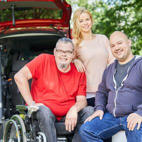 Drei lächelnde Menschen vor dem geöffnetem Kofferraum eines Transporters, einer davon sitzt im Rollstuhl