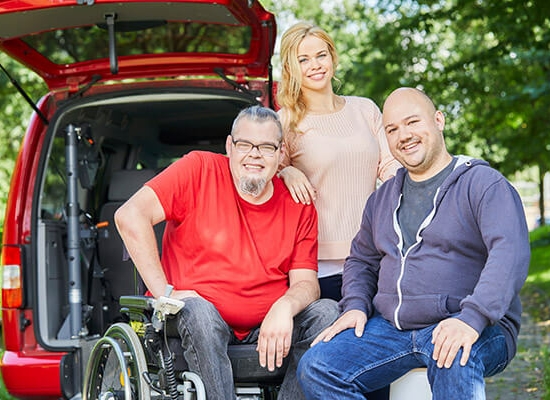 Drei lächelnde Menschen vor dem geöffnetem Kofferraum eines Transporters, einer davon sitzt im Rollstuhl